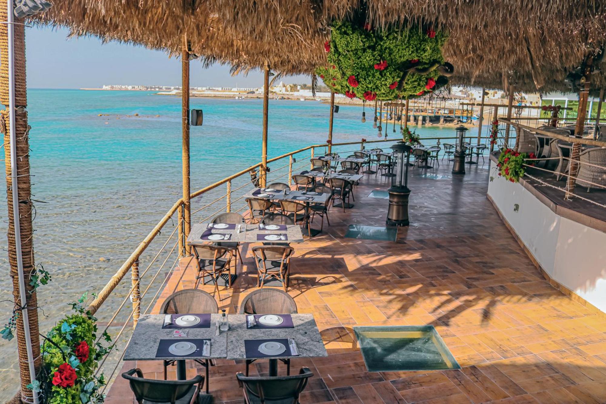Lagoona Beach Luxury Resort And Spa Al-Manamah Bagian luar foto The photo shows a seaside restaurant with a spacious outdoor dining area. It features several tables set with plates and utensils, surrounded by lush greenery and decorative plants. The flooring is made of brown tiles, and there are thatched roofs pr