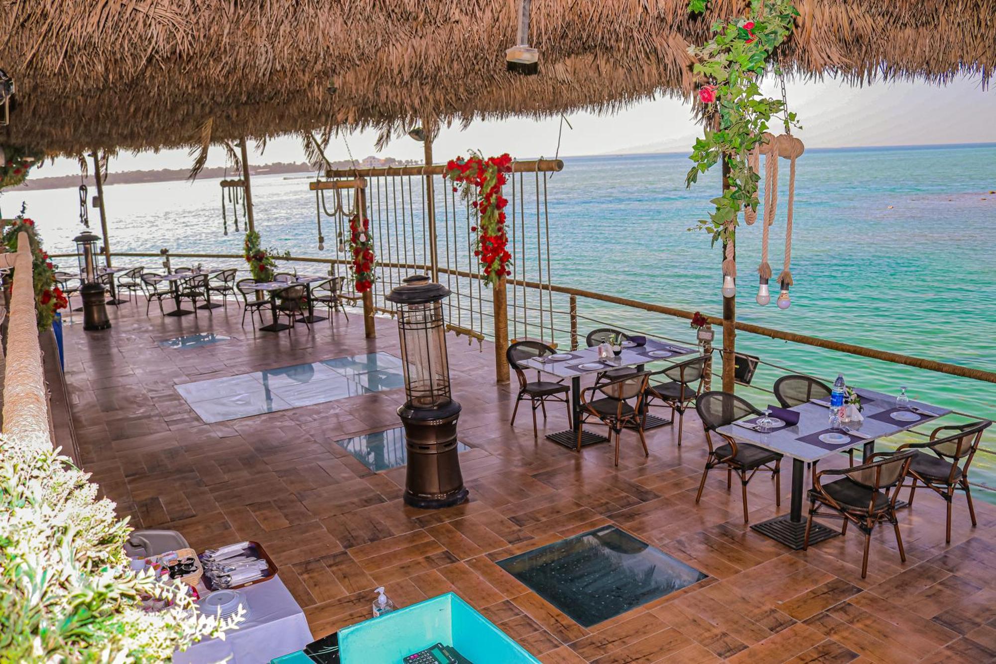 Lagoona Beach Luxury Resort And Spa Al-Manamah Bagian luar foto The photo depicts a coastal restaurant or dining area with an open-air design. It features a wooden floor and tables set for dining, surrounded by chairs. The space is partially covered by a thatched roof, providing shade while allowing for a view of