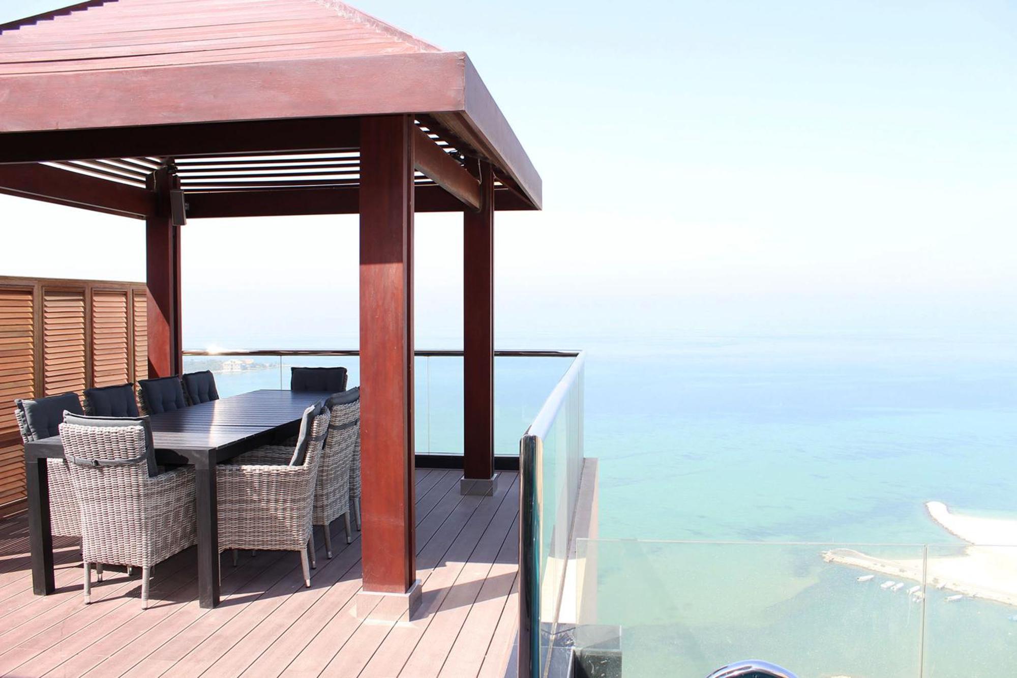 Lagoona Beach Luxury Resort And Spa Al-Manamah Bagian luar foto The photo showcases a stylish outdoor patio or terrace area. It features a wooden structure with a sloped roof, providing a shaded space. There is a large table made of dark wood, surrounded by several wicker chairs. The patio is elevated and has gla