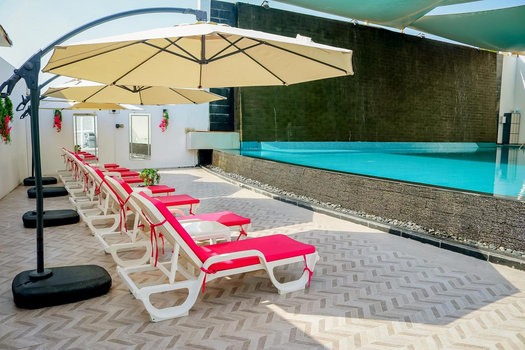 Lagoona Beach Luxury Resort And Spa Al-Manamah Bagian luar foto The photo shows a sunny poolside area with several lounge chairs arranged in a row. Each chair features red cushions and is positioned under large, beige umbrellas for shade. In the background, there is a beautiful swimming pool with clear blue water