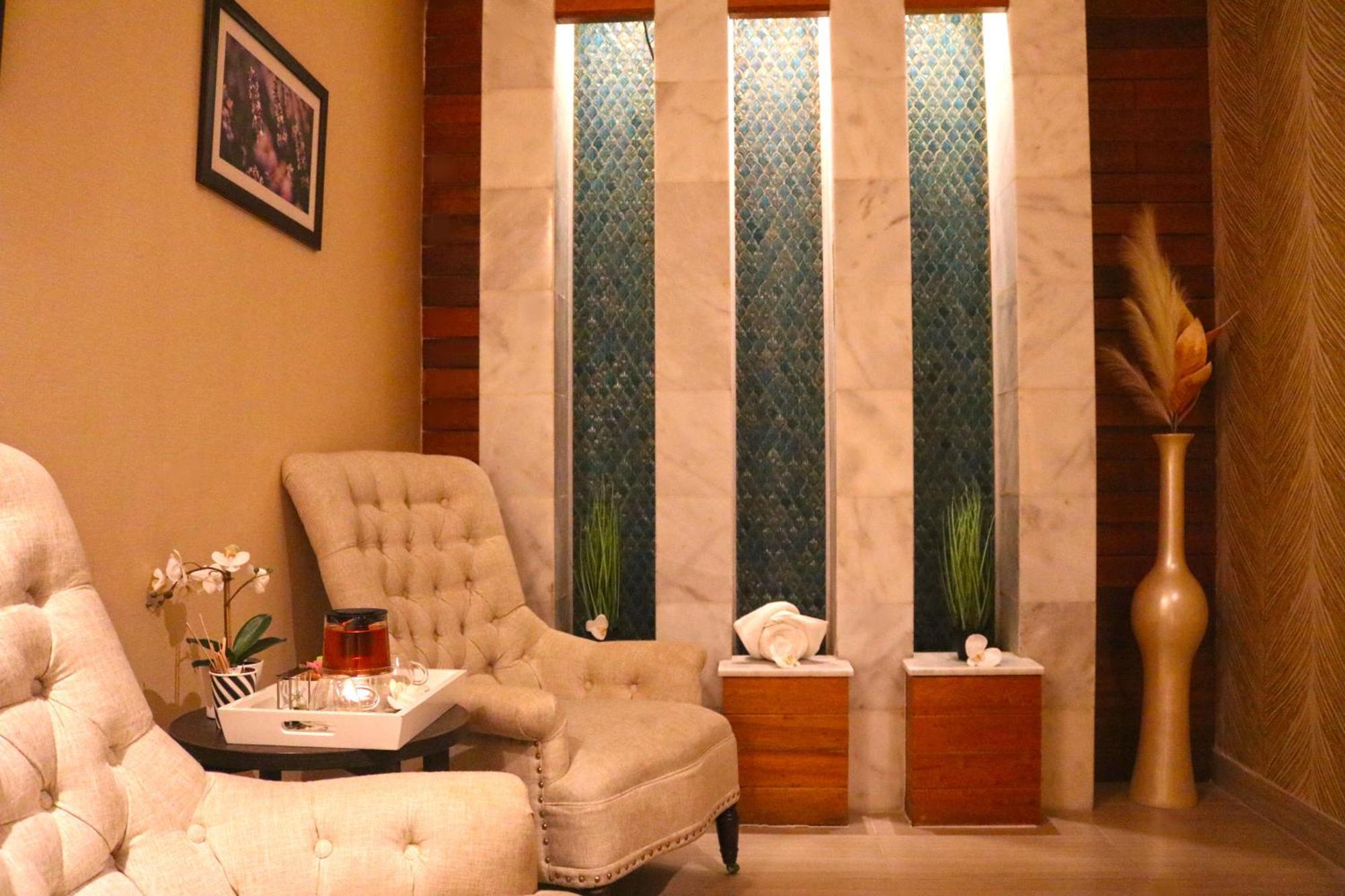 Lagoona Beach Luxury Resort And Spa Al-Manamah Bagian luar foto The image shows a relaxing interior space, likely a waiting area or lounge. There are two plush, beige armchairs positioned near a small table, which holds a glass pitcher and a cup. The background features tall, elegant windows with decorative blue 
