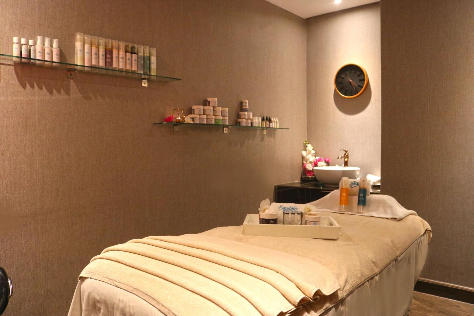 Lagoona Beach Luxury Resort And Spa Al-Manamah Bagian luar foto The photo shows a spa treatment room. There is a massage table draped with a light-colored blanket, creating a relaxing atmosphere. On the wall, there are shelves displaying various skincare products in bottles and jars. A clock is mounted on the wal