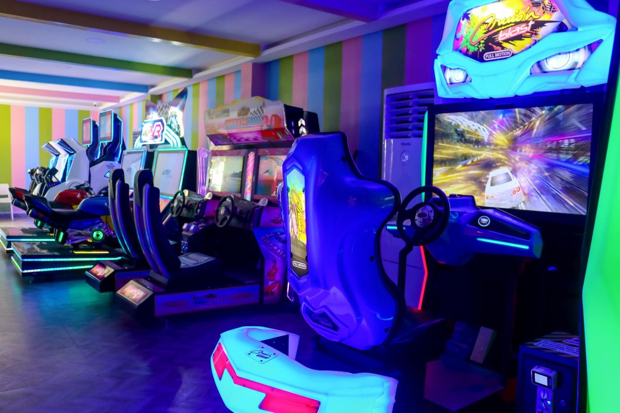 Lagoona Beach Luxury Resort And Spa Al-Manamah Bagian luar foto The photo shows an arcade area filled with various arcade machines. The setting is illuminated with colorful neon lights, creating a vibrant atmosphere. There are several racing game machines with steering wheels and seats, each accompanied by screen