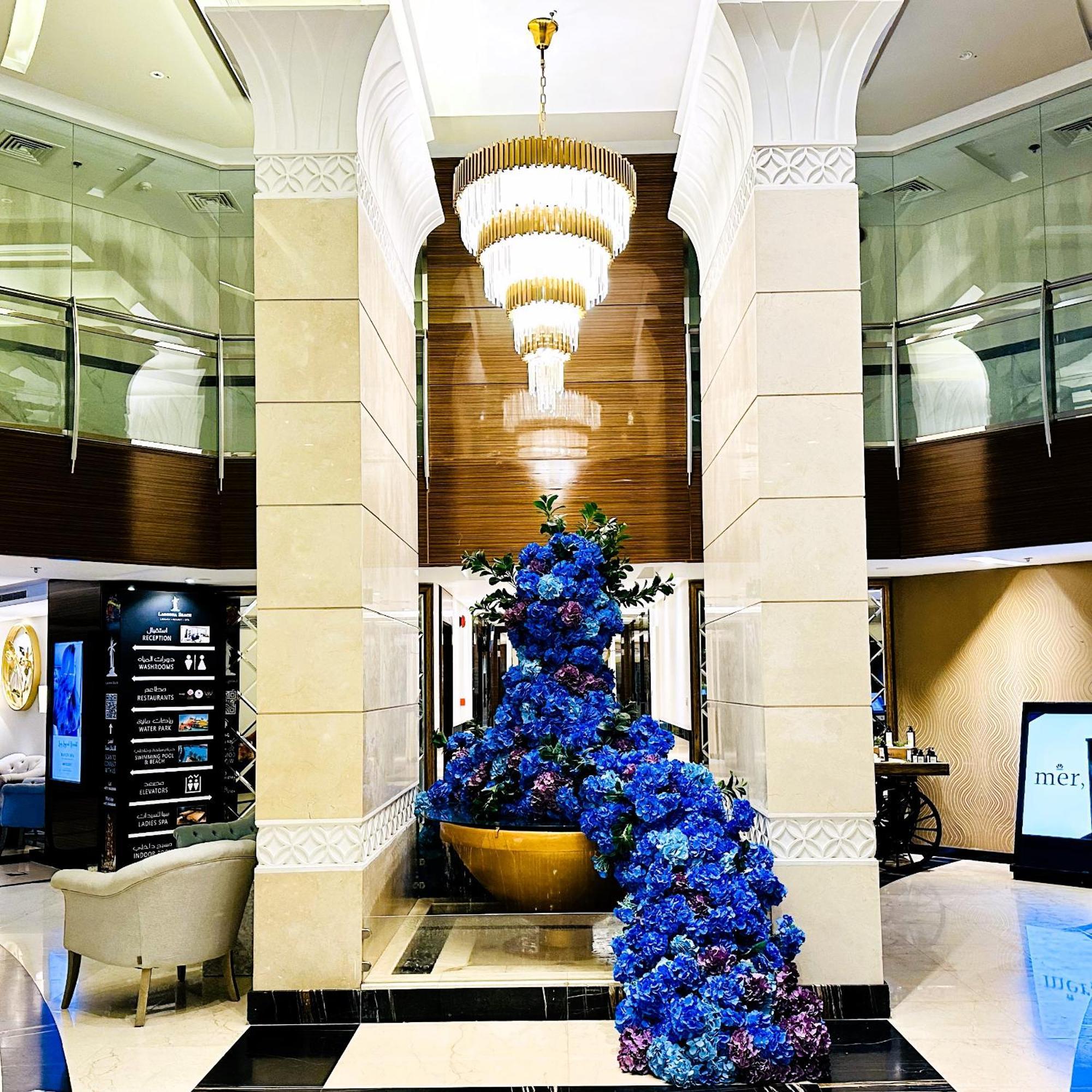 Lagoona Beach Luxury Resort And Spa Al-Manamah Bagian luar foto The photo depicts an elegant lobby or entrance area of a building. Central to the scene is a large floral arrangement featuring vibrant blue flowers. This arrangement is positioned in a decorative bowl. Above it hangs a stylish chandelier with a laye