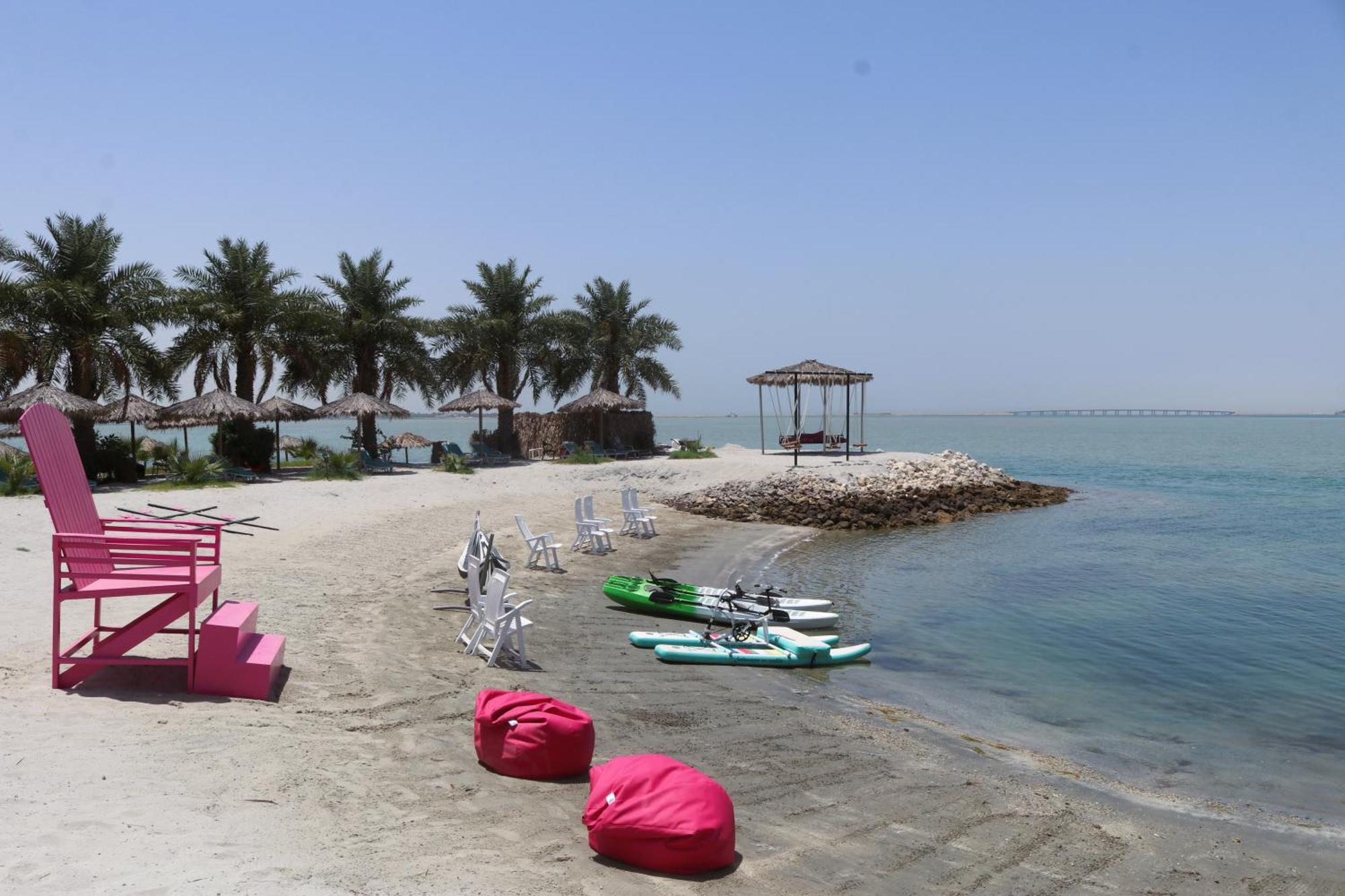 Lagoona Beach Luxury Resort And Spa Al-Manamah Bagian luar foto The photo depicts a serene beach scene with clear blue water and a sunny sky. In the foreground, there are colorful pink bean bags and a large pink chair on the sand. A few white chairs and loungers are positioned near the water's edge. To the side, 