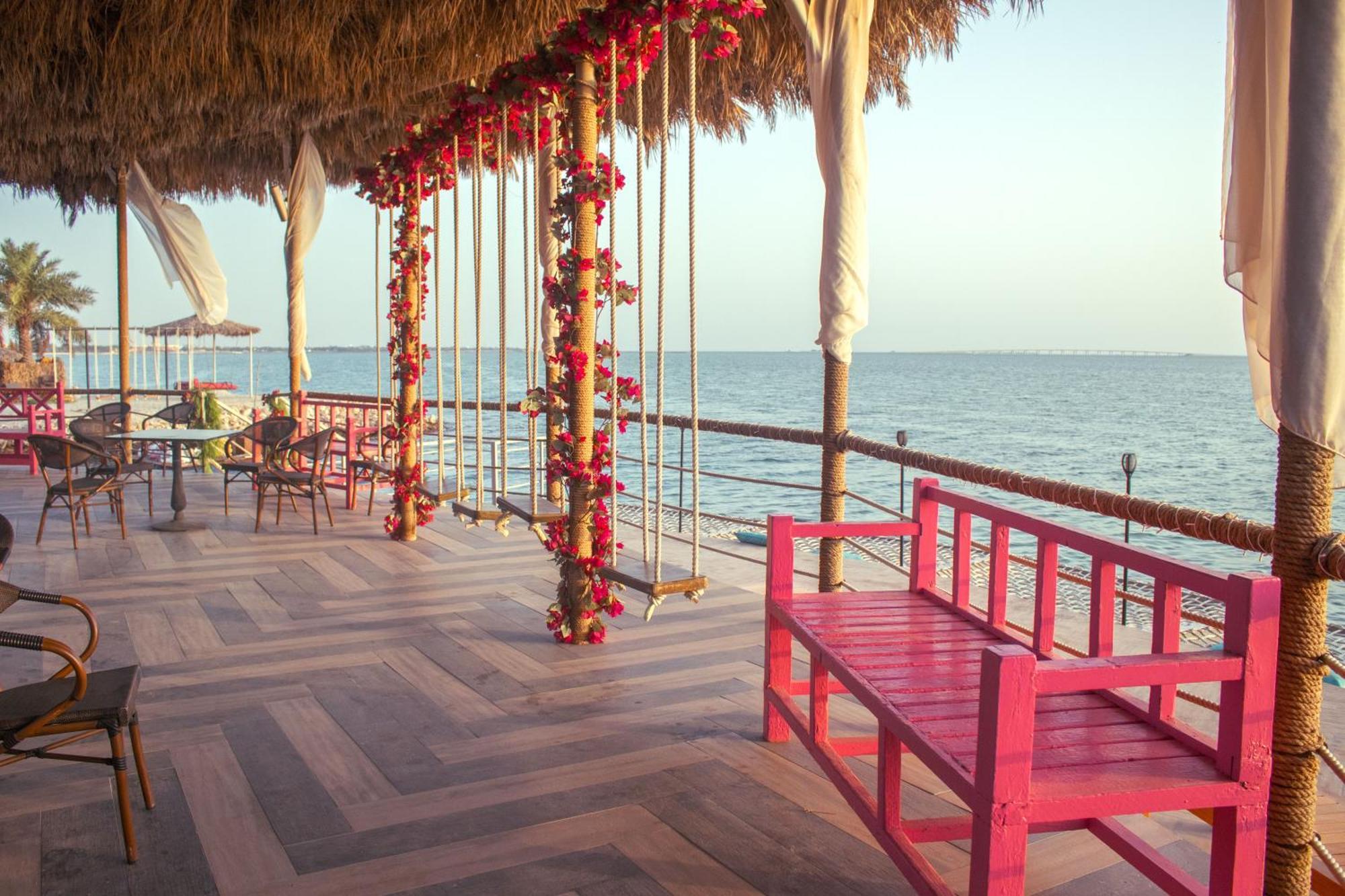 Lagoona Beach Luxury Resort And Spa Al-Manamah Bagian luar foto The photo depicts a scenic outdoor area by the water, with a wooden deck and a thatched roof. Two swings adorned with flowers are hanging from the ceiling, providing a charming touch to the space. In the foreground, there is a bright pink bench, addi
