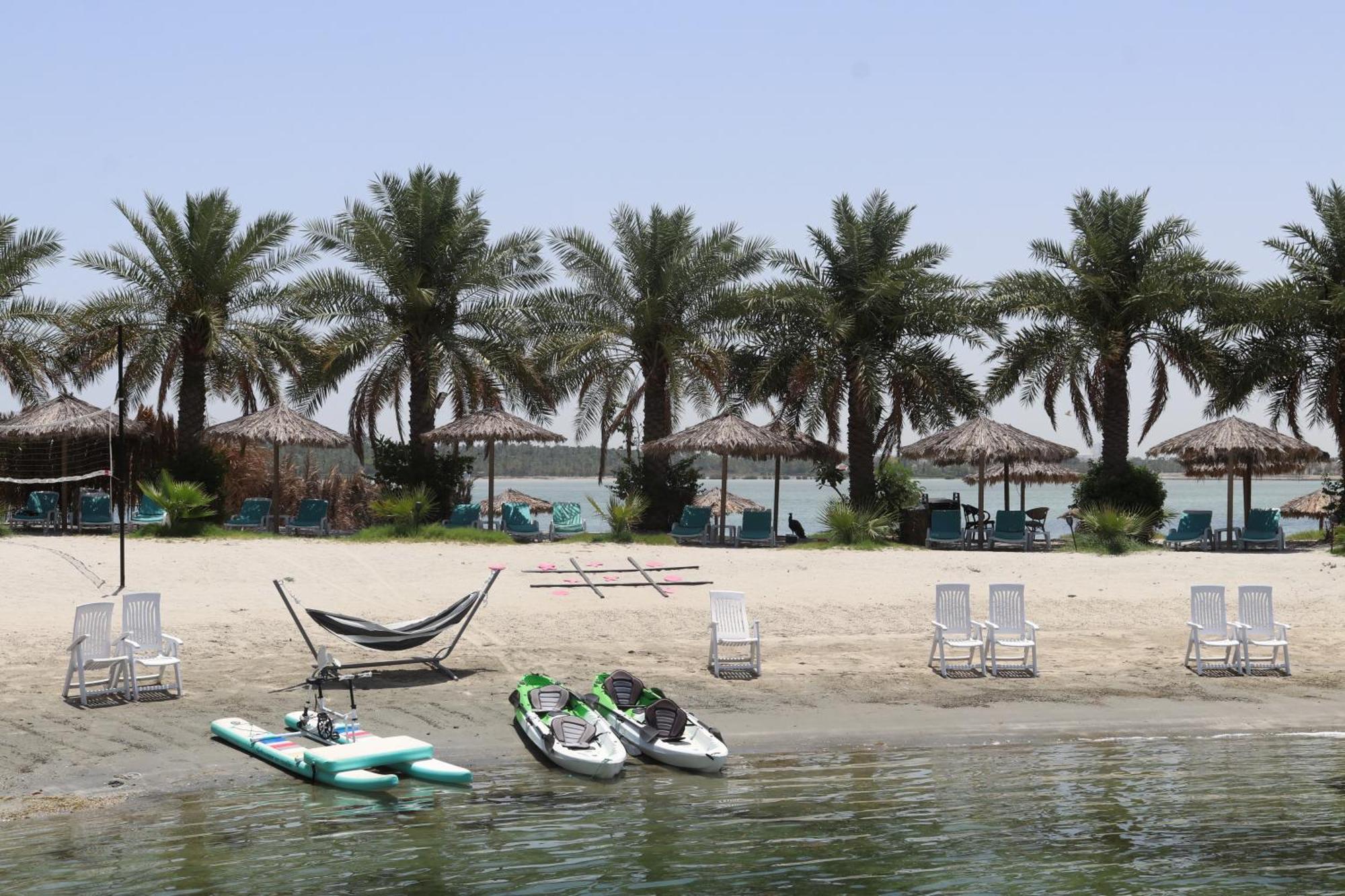 Lagoona Beach Luxury Resort And Spa Al-Manamah Bagian luar foto The photo depicts a serene beach scene with several palm trees lining the shore. There are thatched umbrellas providing shade, and a few lounge chairs are positioned on the sand. A hammock is set up between two supports, inviting relaxation. In the f