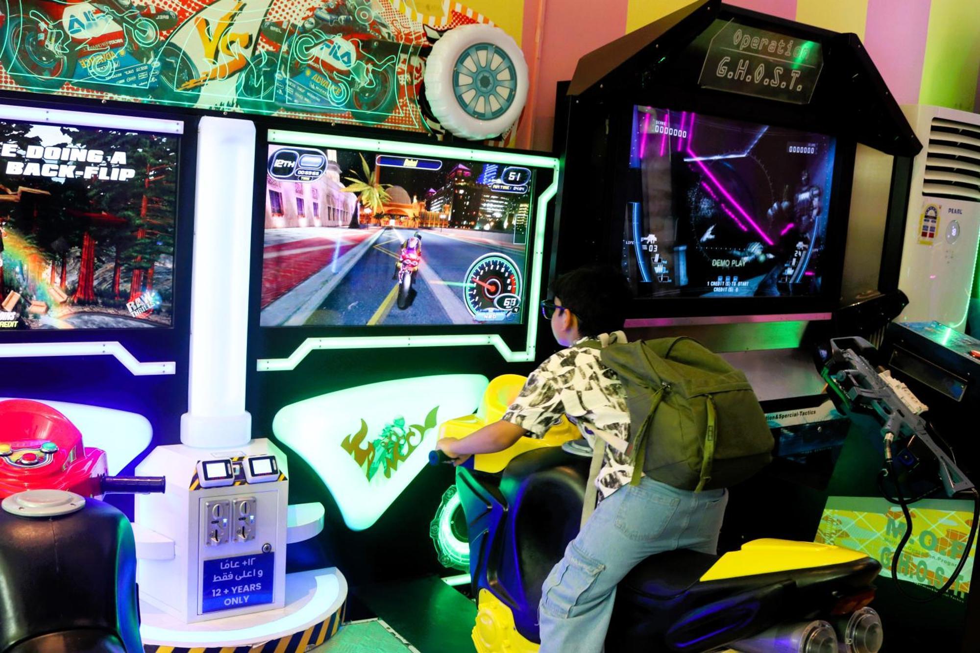 Lagoona Beach Luxury Resort And Spa Al-Manamah Bagian luar foto The photo shows a person playing an arcade motorcycle racing game. They are seated on a motorcycle-shaped seat, actively engaging with the game displayed on the screen in front of them. The screen shows a vibrant racing scene with streets and colorfu