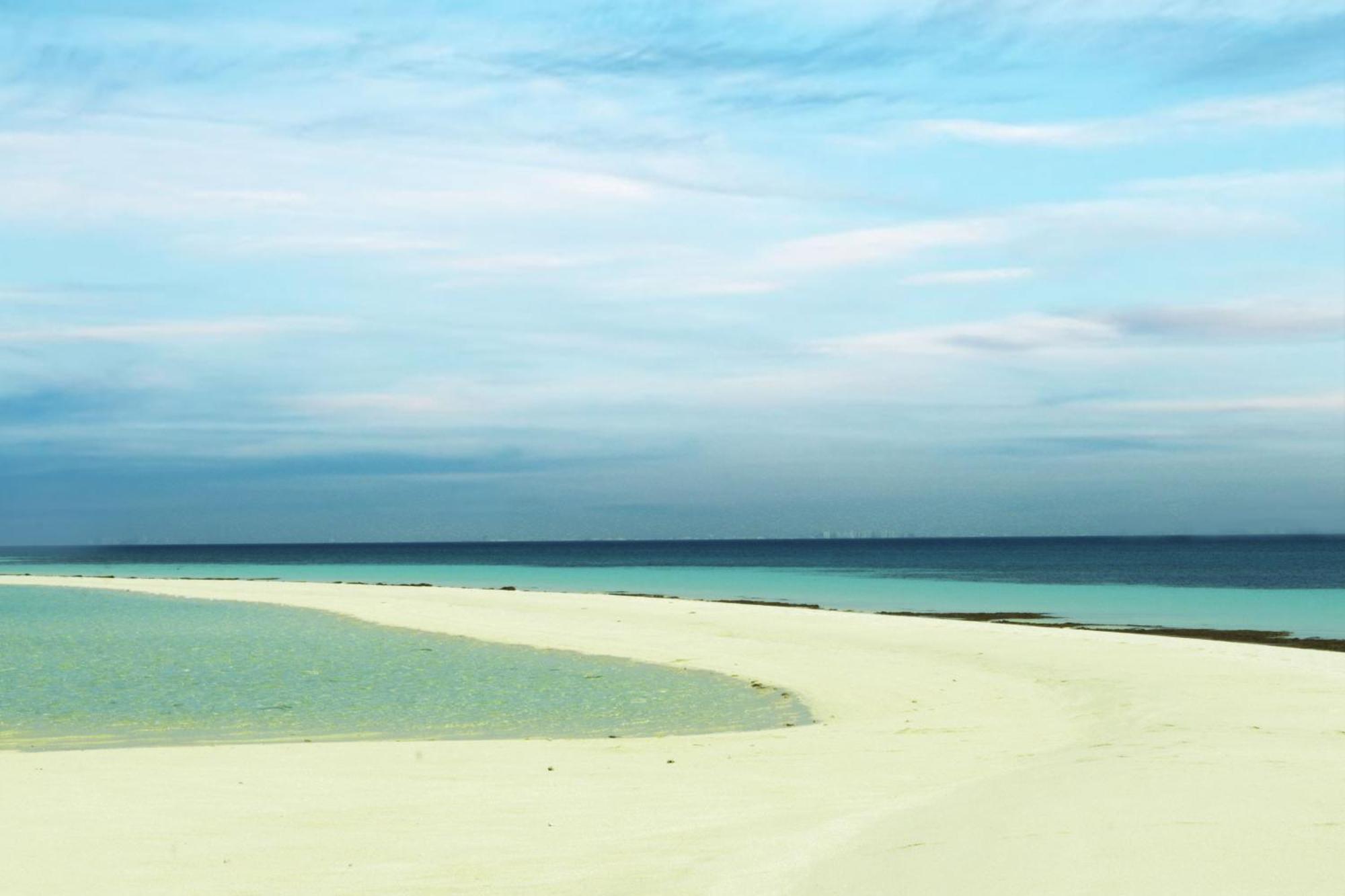 Lagoona Beach Luxury Resort And Spa Al-Manamah Bagian luar foto The photo depicts a serene beach scene with a gentle curve of sandy shore leading into shallow turquoise waters. The calm ocean stretches into the horizon, meeting a sky filled with soft clouds in various shades of blue. The overall atmosphere is tra