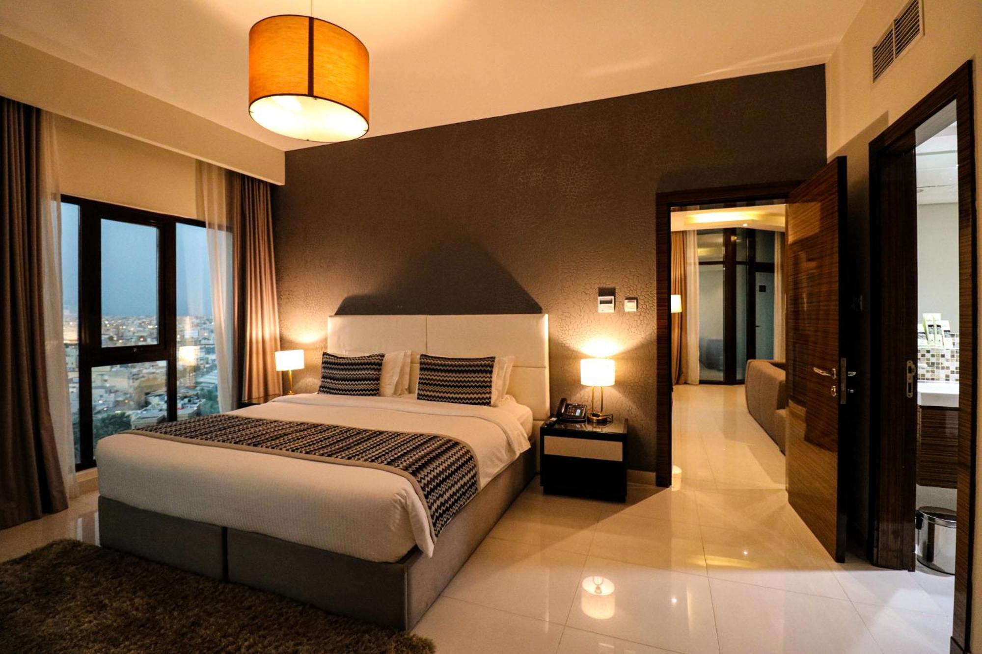 Lagoona Beach Luxury Resort And Spa Al-Manamah Bagian luar foto The photo depicts a modern hotel room or suite. It features a large bed with a white and gray color scheme, accented with decorative pillows. On either side of the bed, there are sleek bedside lamps providing warm lighting. The walls are a dark color