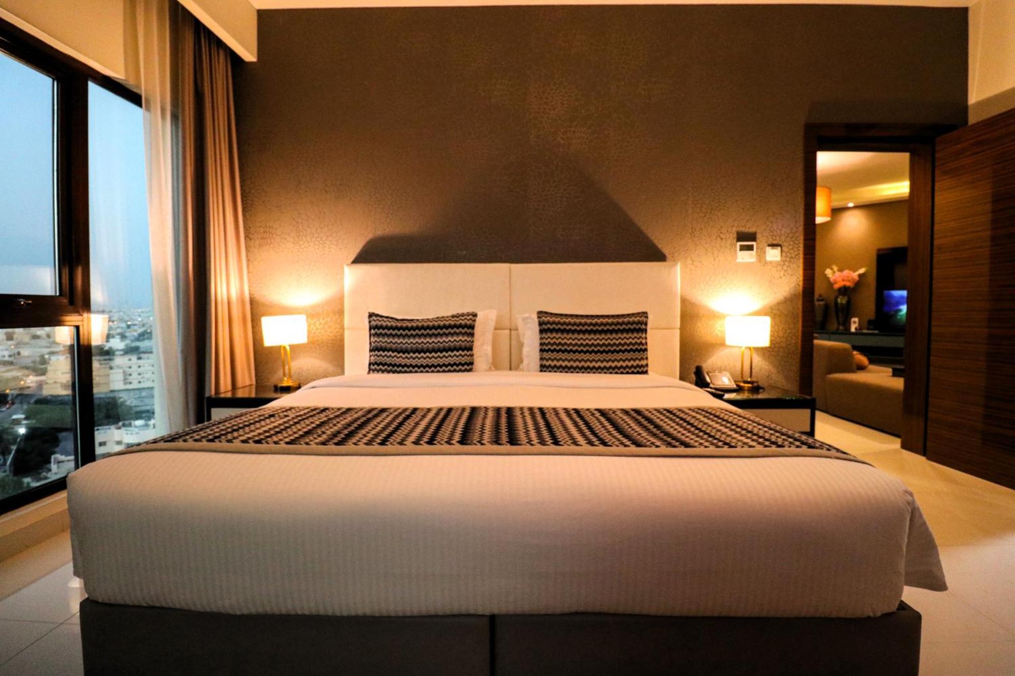 Lagoona Beach Luxury Resort And Spa Al-Manamah Bagian luar foto The photo depicts a modern hotel room featuring a large bed with a white comforter and two decorative pillows. The headboard is light-colored, and the walls are painted a dark hue. On either side of the bed, there are bedside tables with lamps. The r