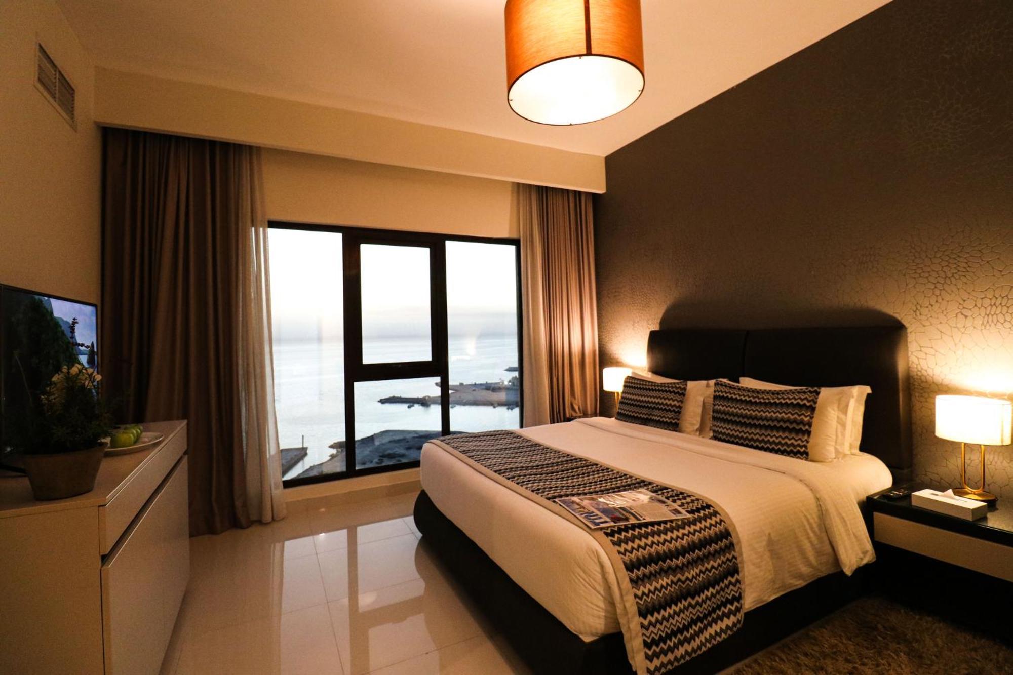 Lagoona Beach Luxury Resort And Spa Al-Manamah Bagian luar foto The photo shows a modern hotel room featuring a neatly made bed with decorative pillows. The room has a stylish design with a dark accent wall and a large window that offers a view of the outdoors. Natural light comes in, giving the space a bright an