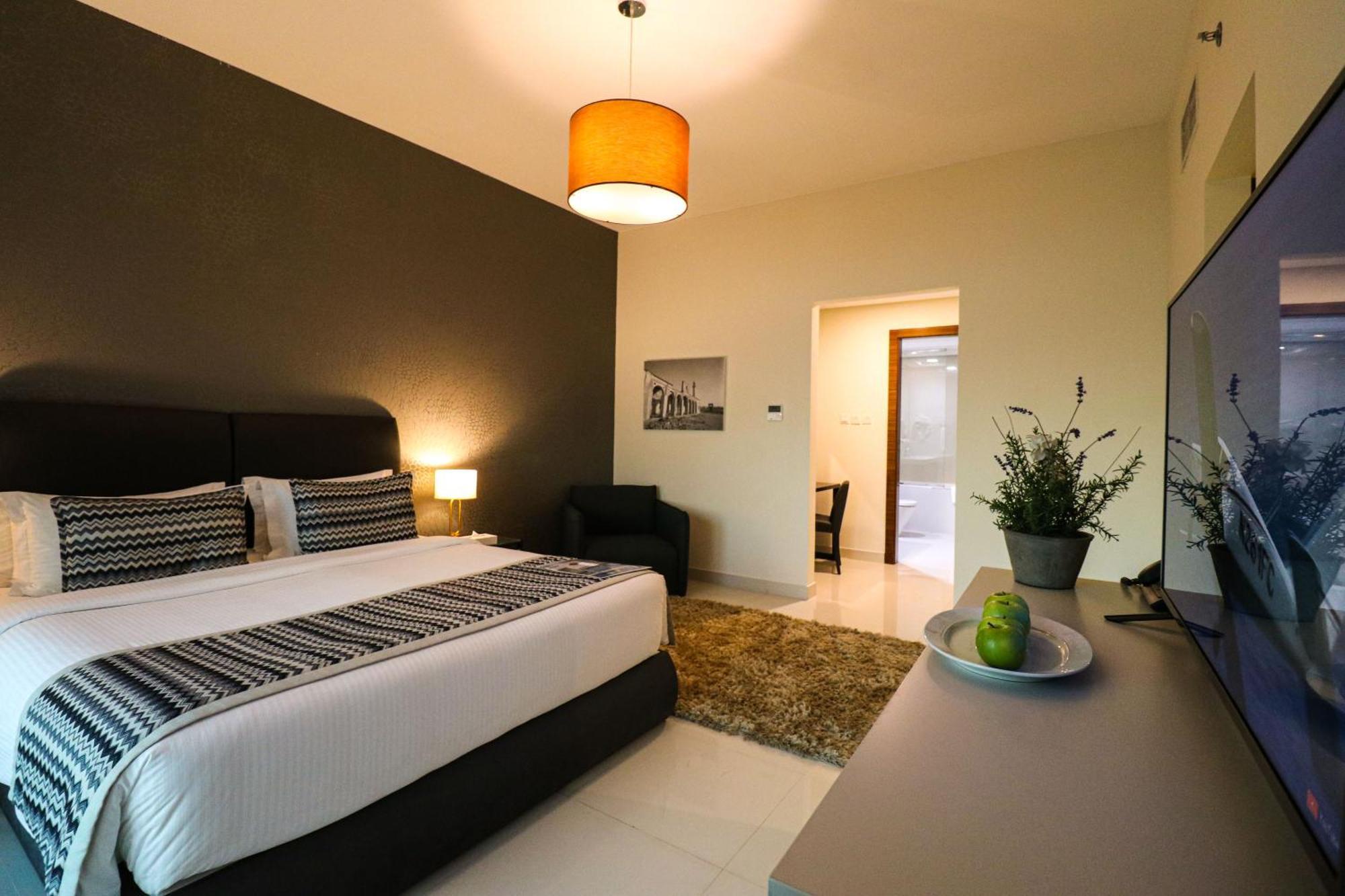 Lagoona Beach Luxury Resort And Spa Al-Manamah Bagian luar foto The photo shows a modern hotel room. It features a large bed with a neatly made white and dark-colored duvet. On either side of the bed, there are bedside tables with lamps. The wall behind the bed is painted in a dark color, creating a contrast with