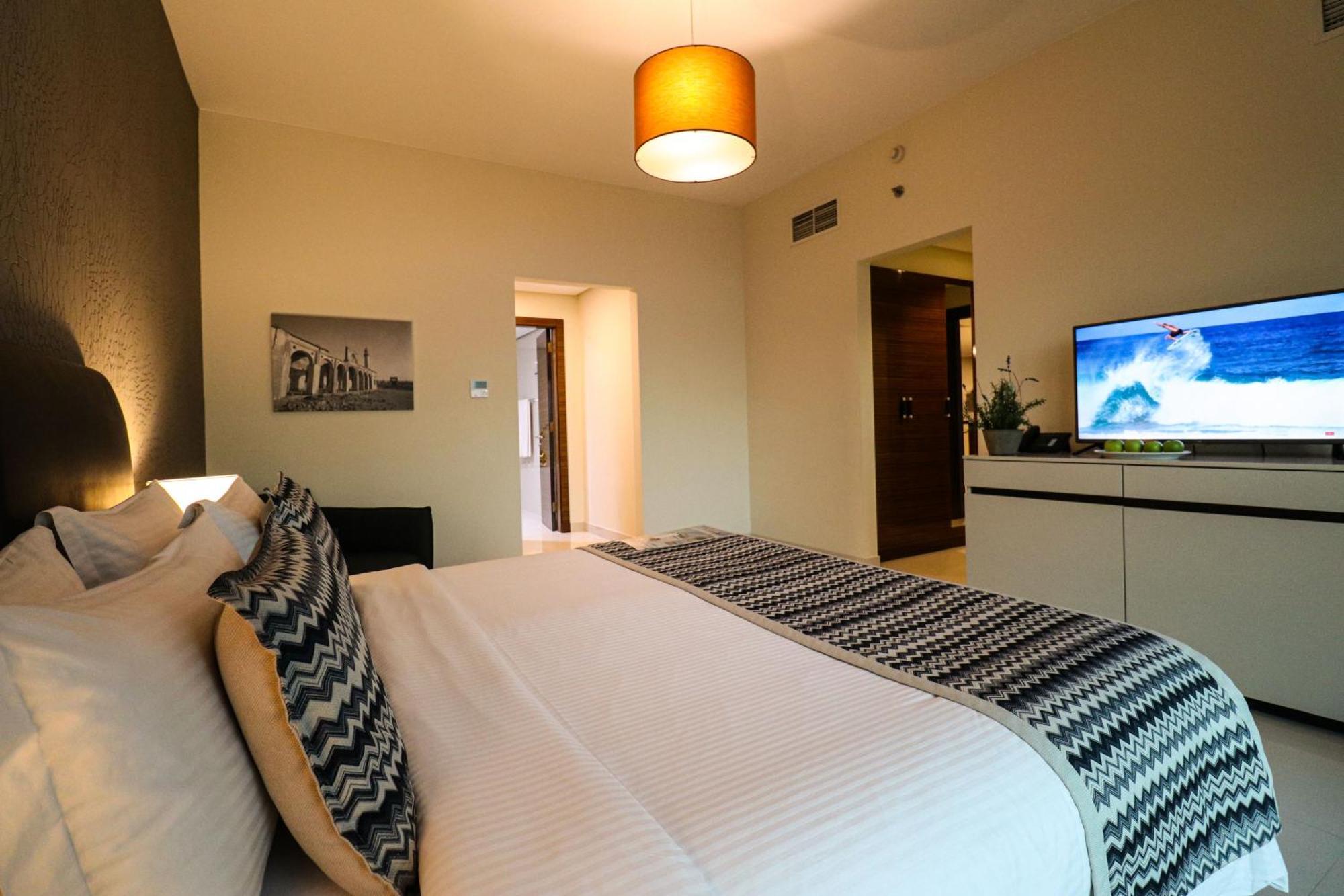 Lagoona Beach Luxury Resort And Spa Al-Manamah Bagian luar foto The photo shows a well-decorated hotel room. There is a large bed with white linens and decorative pillows featuring a zigzag pattern. On the wall beside the bed is a piece of artwork, and there is a modern television on a white furniture unit opposi
