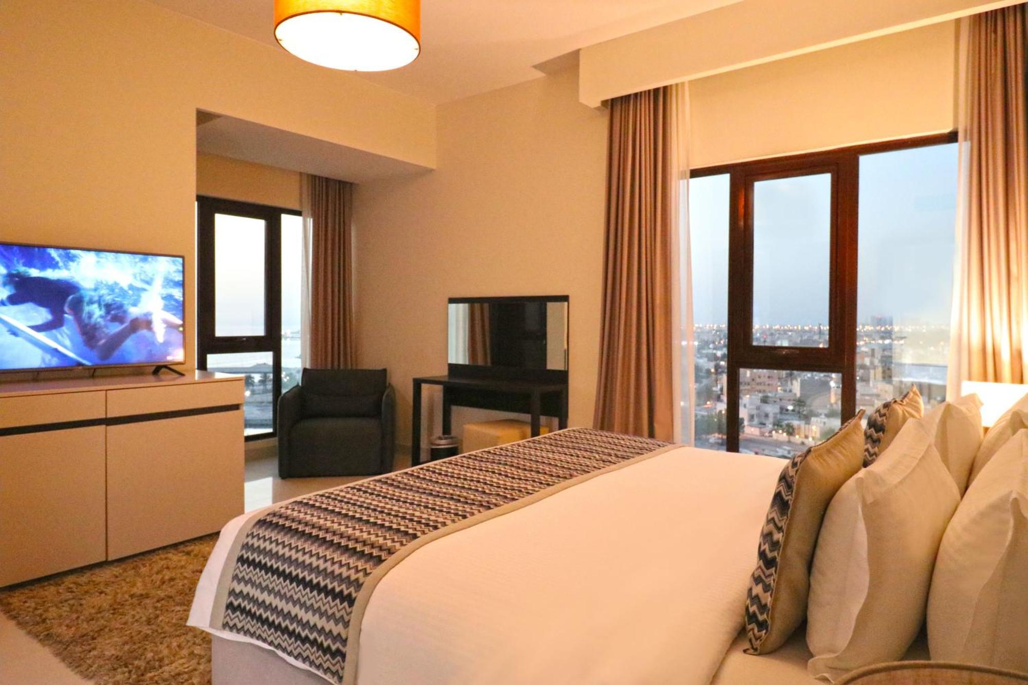 Lagoona Beach Luxury Resort And Spa Al-Manamah Bagian luar foto The photo shows a modern hotel room or suite. It features a neatly made bed with decorative pillows in the foreground. To the left, there is a TV on a cabinet, and further back, a comfortable chair is situated in front of a large window that likely o
