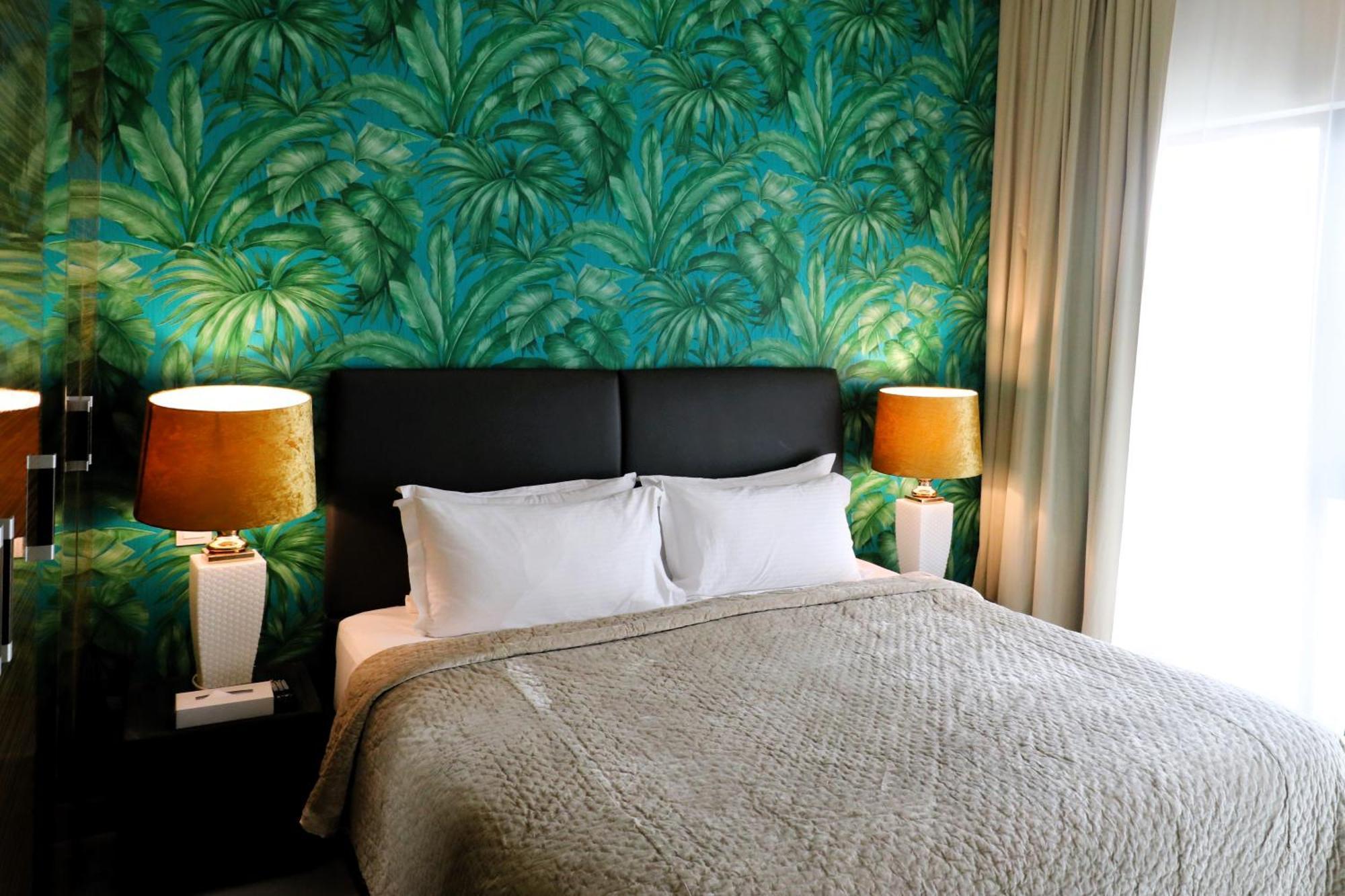 Lagoona Beach Luxury Resort And Spa Al-Manamah Bagian luar foto The photo shows a stylish bedroom featuring a large bed with white pillows and a gray blanket. The headboard is upholstered in black, providing a modern touch. Behind the bed, there is a vibrant green wallpaper with a tropical leaf pattern, adding a 