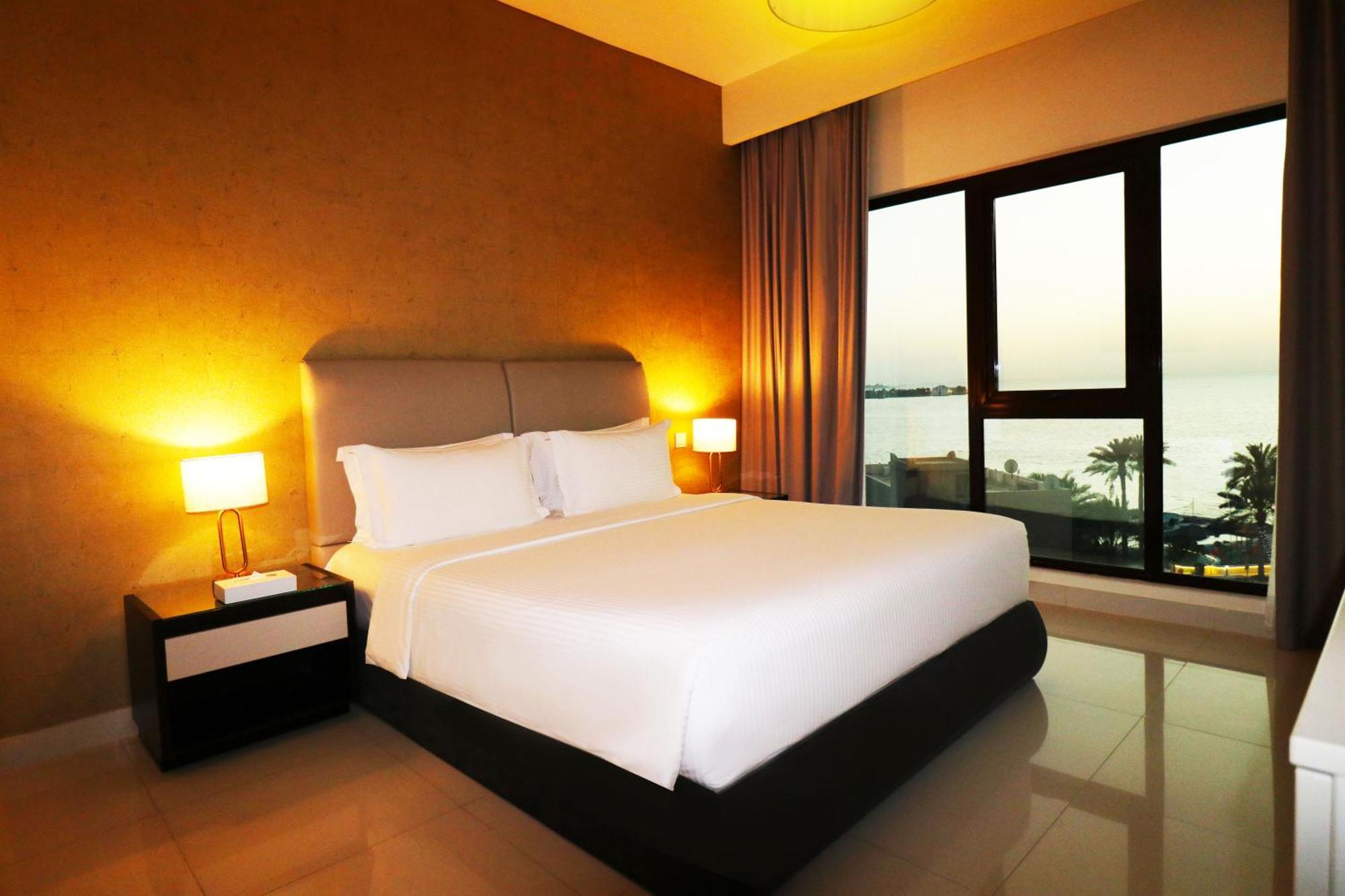 Lagoona Beach Luxury Resort And Spa Al-Manamah Bagian luar foto The photo shows a modern hotel bedroom with a large, comfortable bed dressed in white linens. The room features warm lighting, including bedside lamps, creating a cozy atmosphere. There are large windows with a view of the outdoors, possibly overlook