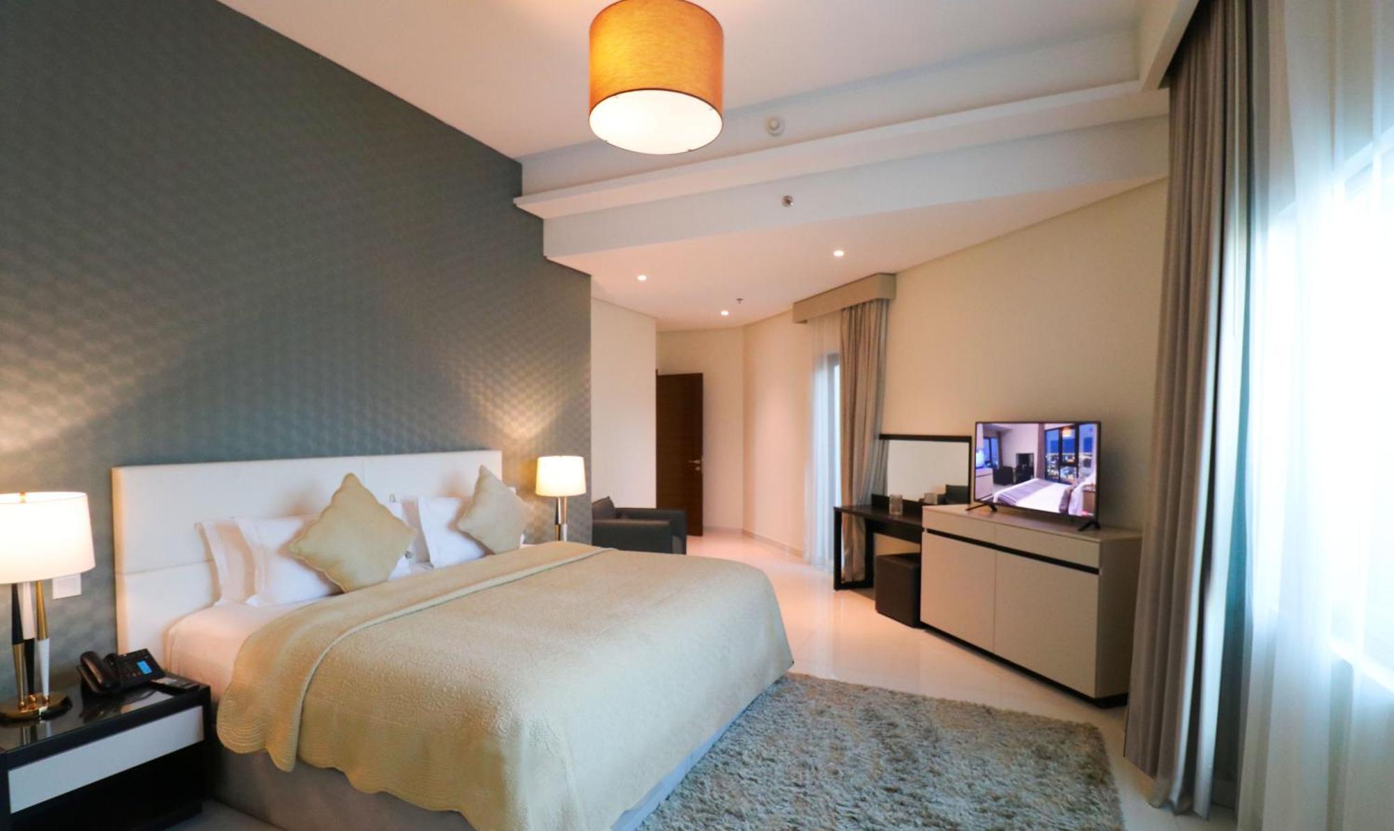 Lagoona Beach Luxury Resort And Spa Al-Manamah Bagian luar foto The photo shows a modern hotel room featuring a large bed with a light-colored blanket and decorative pillows. The walls are painted in a subtle shade, and there is a textured accent wall behind the bed. On one side, a bedside lamp provides warm ligh