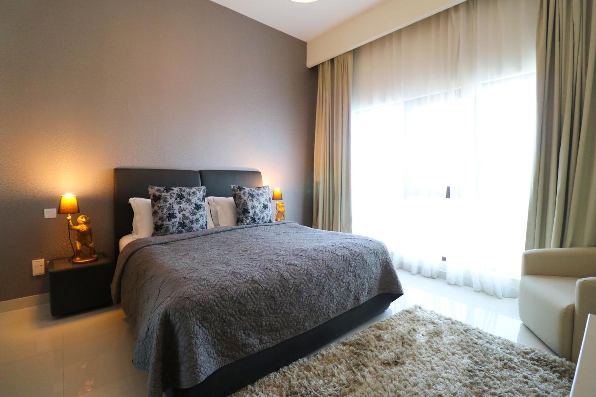 Lagoona Beach Luxury Resort And Spa Al-Manamah Bagian luar foto The photo shows a stylish bedroom with a modern design. It features a large bed with a dark gray bedspread and decorative pillows. On either side of the bed are two wooden nightstands, each with a lamp. There is a large window covered with light, flo